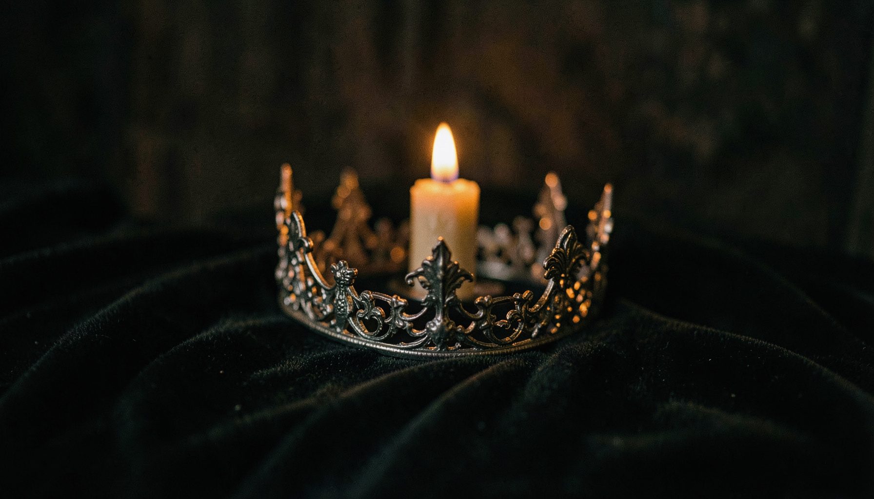 Moody still life of an iron crown resting on dark velvet, lit by a single amber candle.